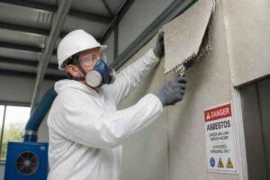 Pracownik w odzieży ochronnej i masce usuwa płytę azbestową ze ściany oznaczonej ostrzeżeniem Danger Asbestos